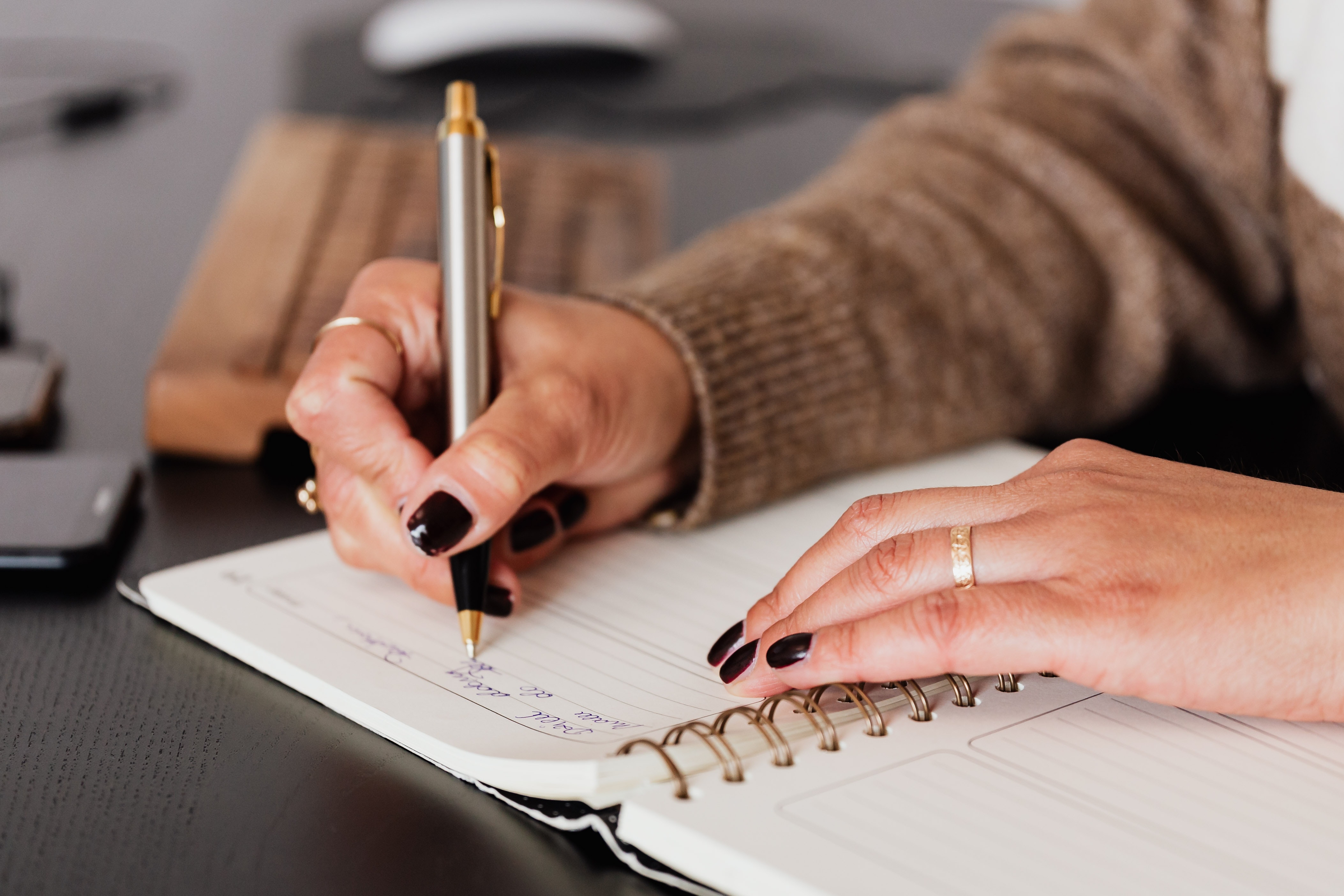 Image of woman with black painted fingernails taking notes in notebook using silver and gold pen.
